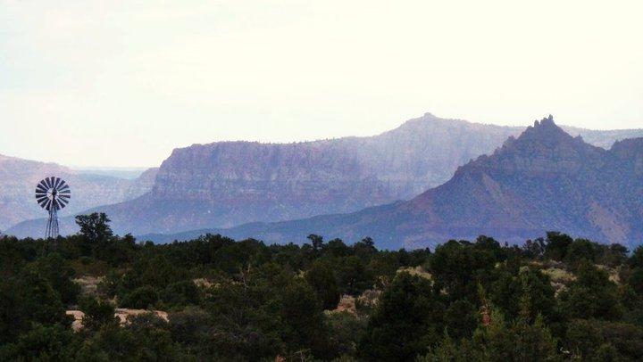 A scenic landscape featuring a windmill in the foreground, surrounded by greenery, with majestic mountain ranges visible in the background under a cloudy sky. Gooseberry Mesa mountain bike trail.
