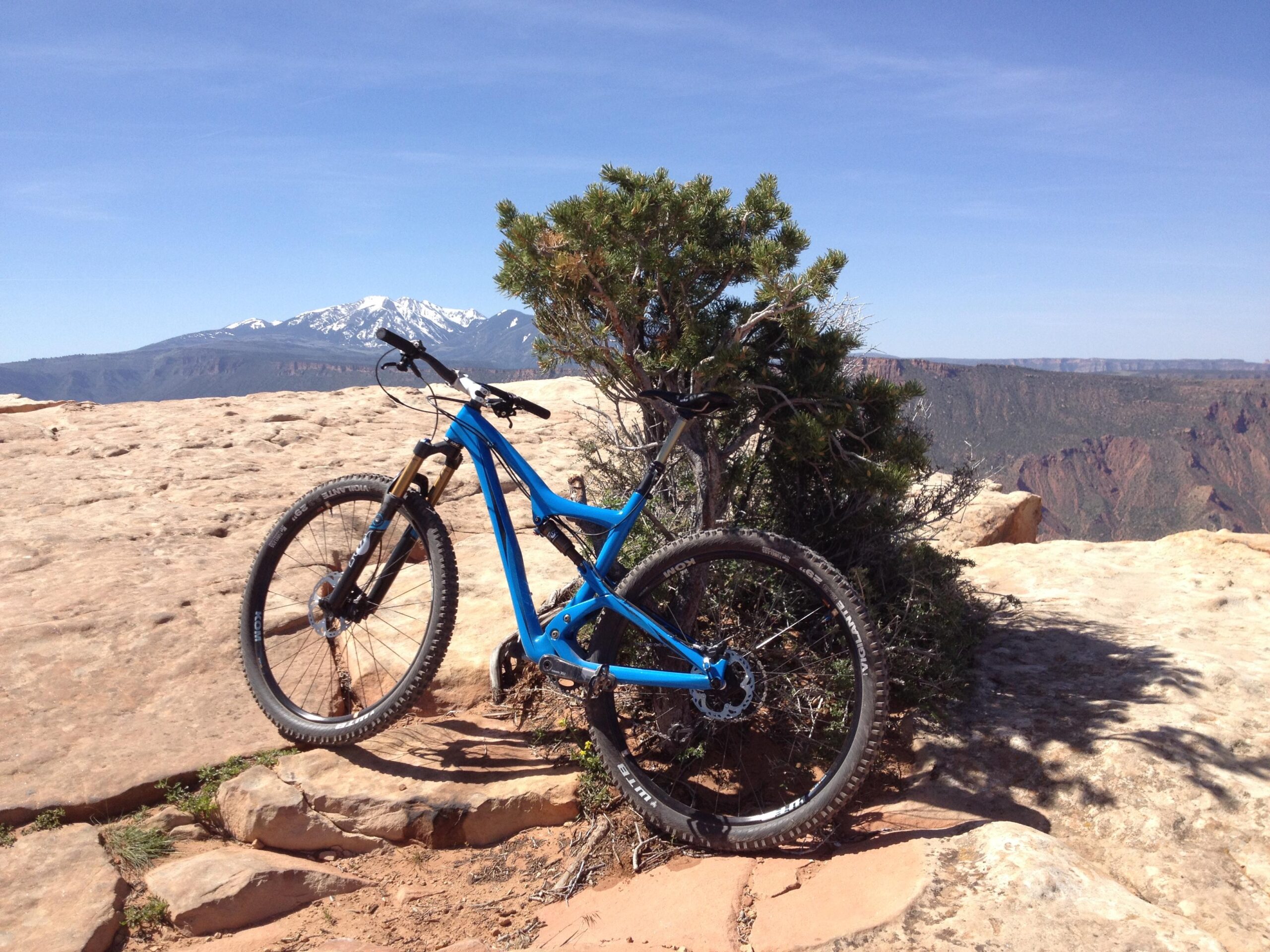 A blue mountain bike leaning against a small shrub on a rocky outcrop, with a vast mountainous landscape and blue sky in the background. Snow-capped peaks are visible in the distance. Top Of The World mountain bike trail.