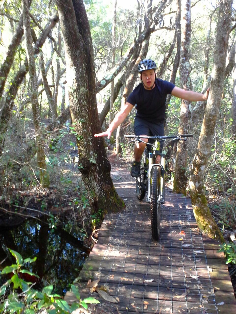 A young man rides a mountain bike on a narrow wooden path surrounded by trees, demonstrating a dynamic pose. He is wearing a helmet and a black shirt, with one arm extended for balance as he navigates the trail. The path runs alongside a reflective body of water, with sunlight filtering through the foliage, creating a vibrant outdoor setting. Longleaf Pine Greenway Trail mountain bike trail.