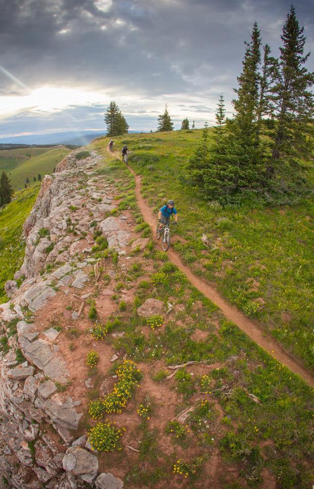 Mountain bikers riding along a rugged dirt trail atop a rocky ridge, surrounded by green grass and wildflowers, with a cloudy sky in the background. Vail Mountain Bike Park mountain bike trail.