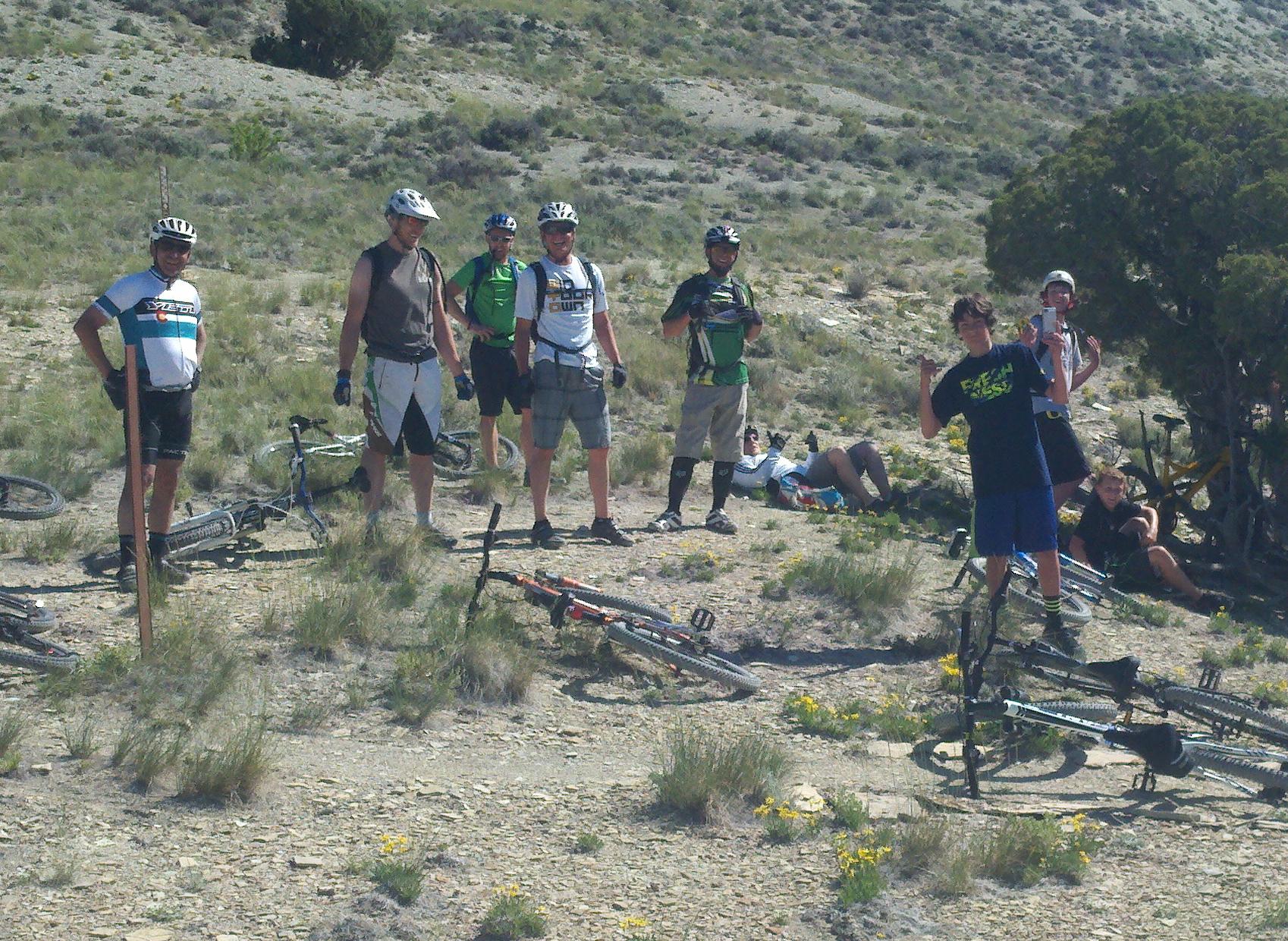 A group of eight mountain bikers gathered on a rugged trail surrounded by shrubs and grass. Some are standing next to their bikes, while others are sitting or reclining on the ground. The riders are wearing various cycling outfits and helmets, showing a mix of casual and sport styles. The sunny, mountainous landscape creates a lively atmosphere. Wilkins Peak Trails mountain bike trail.