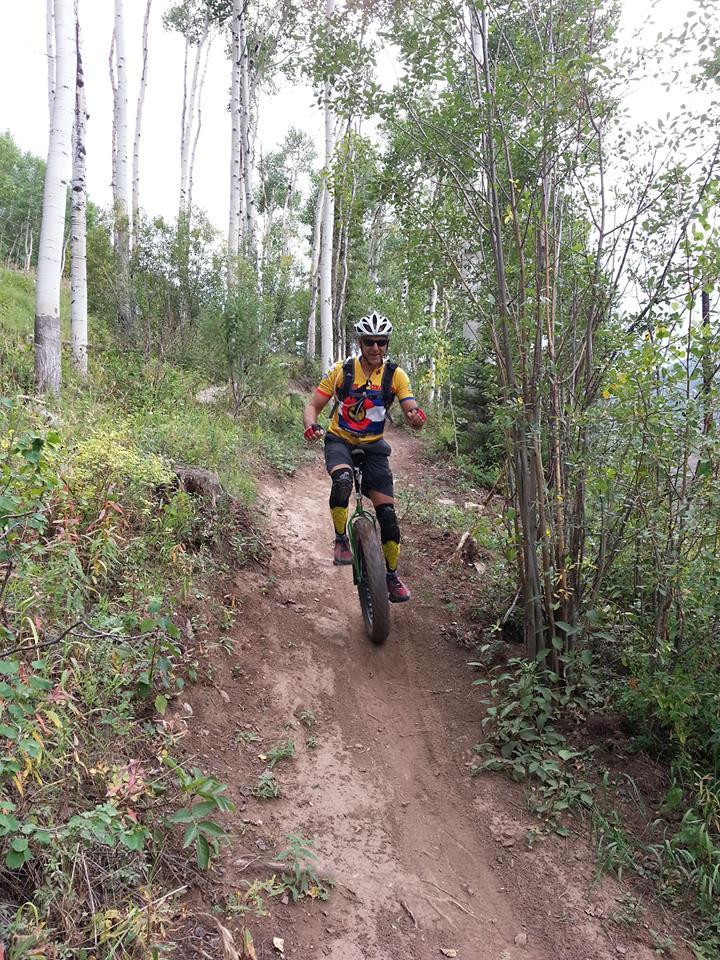 A mountain biker wearing a helmet and protective gear rides along a narrow dirt trail surrounded by tall trees and greenery. The rider is seen actively navigating the trail, showcasing the sport of mountain biking in a natural setting. Vail Mountain Bike Park mountain bike trail.