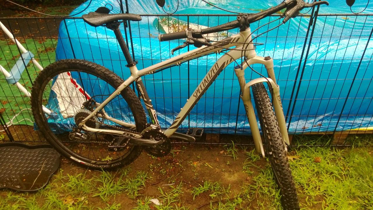Specialized Rockhopper Comp 29: A gray mountain bike leaning against a black fence, with a blue tarp visible in the background. The bike features thick tires and a front suspension fork, suggesting it is designed for off-road use. The ground is grassy and somewhat muddy, indicating recent rain.