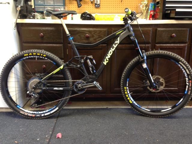 Knolly Chilcotin: A black Knolly mountain bike with yellow and green accents, featuring large tires, a full suspension system, and a gear system. The bike is positioned on a rubber floor, with a wooden workbench in the background displaying tools and other bike accessories.