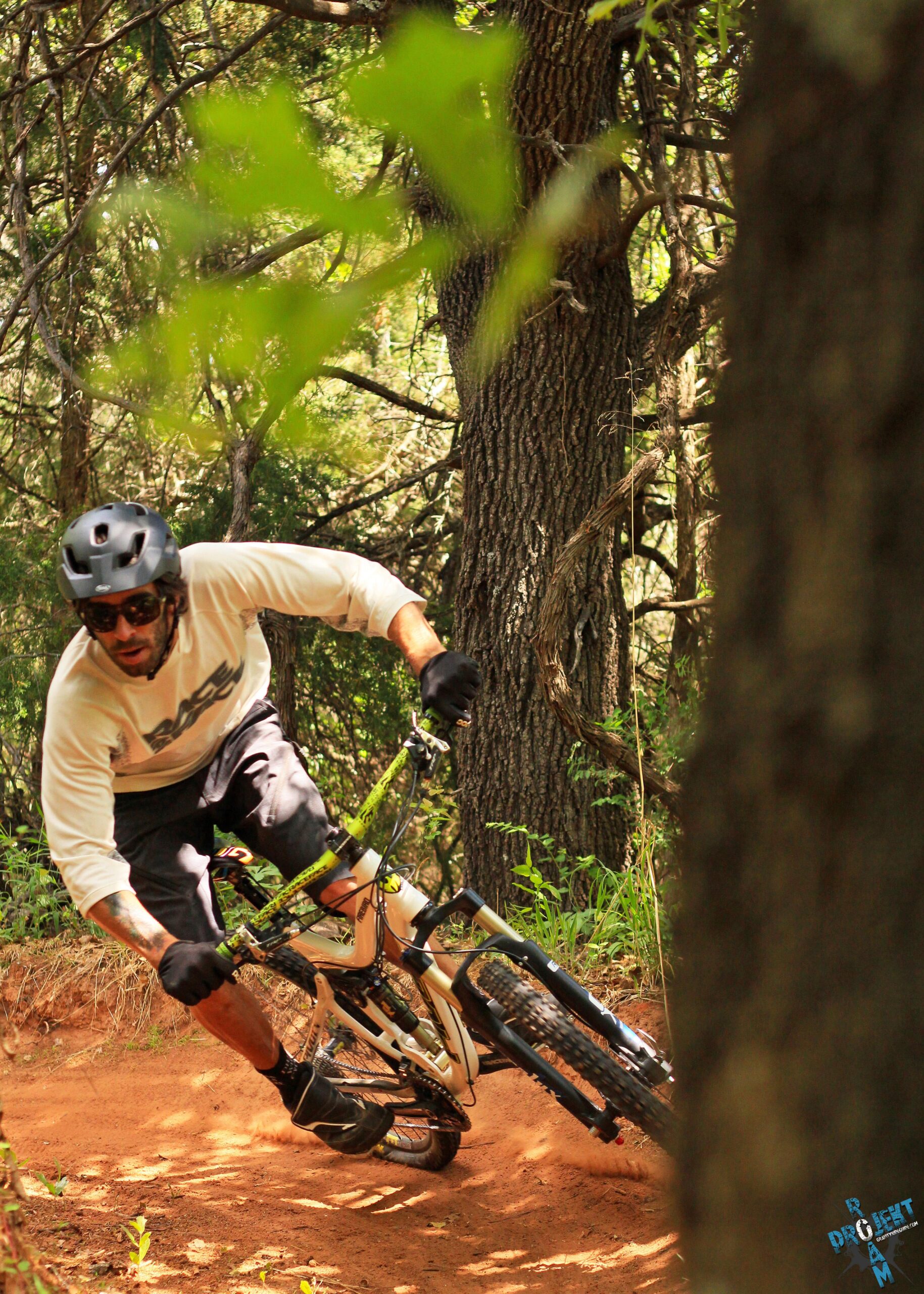 A mountain biker leans into a curve on a dirt trail in a forested area, surrounded by green trees and plants. The rider wears a helmet, sunglasses, and gloves, demonstrating skillful maneuvering on the bike as dust is kicked up from the ground. Lake Stanley Draper mountain bike trail.