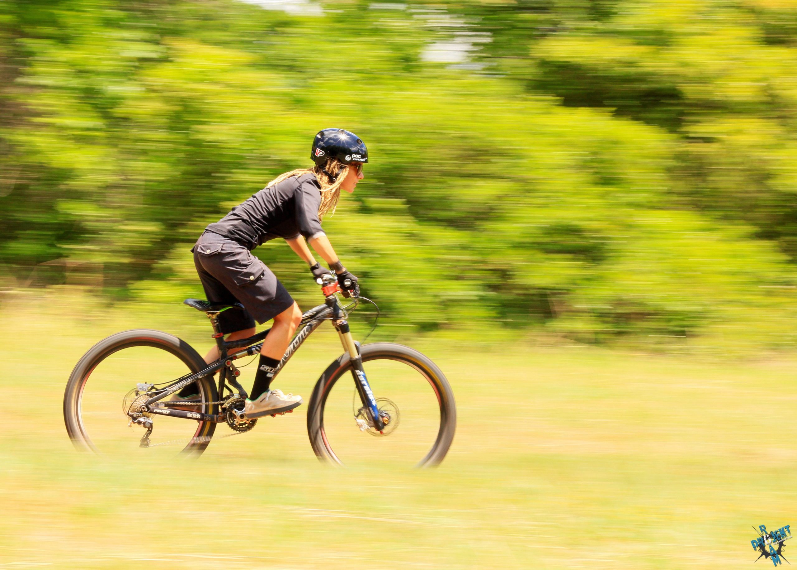 A cyclist in a black helmet and attire rides swiftly on a mountain bike through a grassy area, surrounded by lush greenery. The image captures a sense of motion, highlighted by the blurred background. Lake Stanley Draper mountain bike trail.