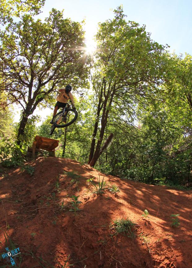 A person riding a mountain bike jumps off a wooden ramp in a forested area, surrounded by green trees and sunlight filtering through the leaves. The biker is mid-air, showcasing a dynamic pose while performing a trick above the sandy terrain. Lake Stanley Draper mountain bike trail.