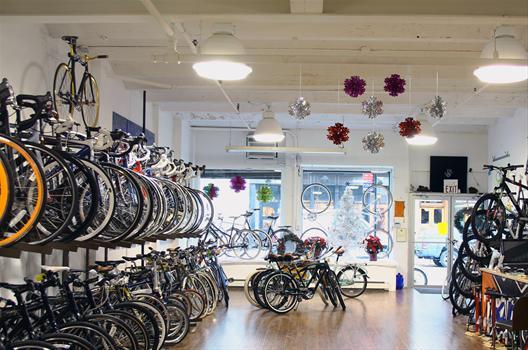 A brightly lit bike shop interior featuring multiple bicycles displayed on walls and in the center. The shop has wooden flooring and decorative elements like colorful ornaments hanging from the ceiling. Large windows allow natural light and provide a view of the outside street.