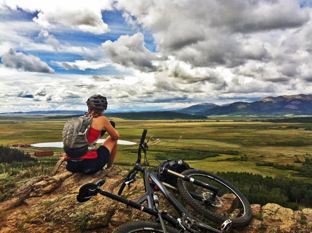 A person sitting on a rocky ledge, overlooking a vast landscape of fields and mountains under a partly cloudy sky. A mountain bike rests beside them, and they are wearing a helmet and a backpack, enjoying the scenic view. Colorado Trail: Kenosha Pass To Breckenridge mountain bike trail.