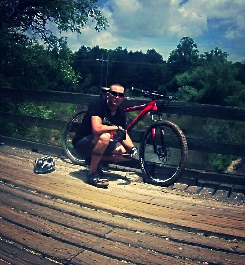 A person squatting next to a red mountain bike on a wooden bridge, wearing sunglasses and gloves, with a bicycle helmet nearby. The background features green trees and a cloudy blue sky. Dawson Forest mountain bike trail.
