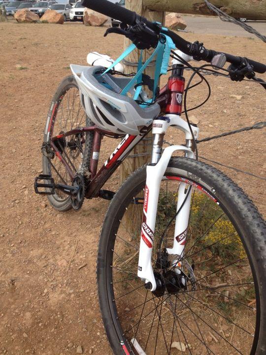 A mountain bike with a white and red front fork, parked next to a wooden post in an outdoor area. A white helmet is attached to the bike. The ground is dry and rocky, with parked cars visible in the background.
