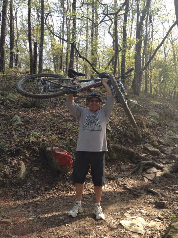 Specialized Hardrock Comp Disc: A cyclist in a gray t-shirt and helmet stands on a dirt path in a forest, joyfully holding their mountain bike above their head. Surrounding trees are beginning to show signs of spring with fresh green leaves. The terrain is rocky, and sunlight filters through the branches, creating a vibrant outdoor scene.