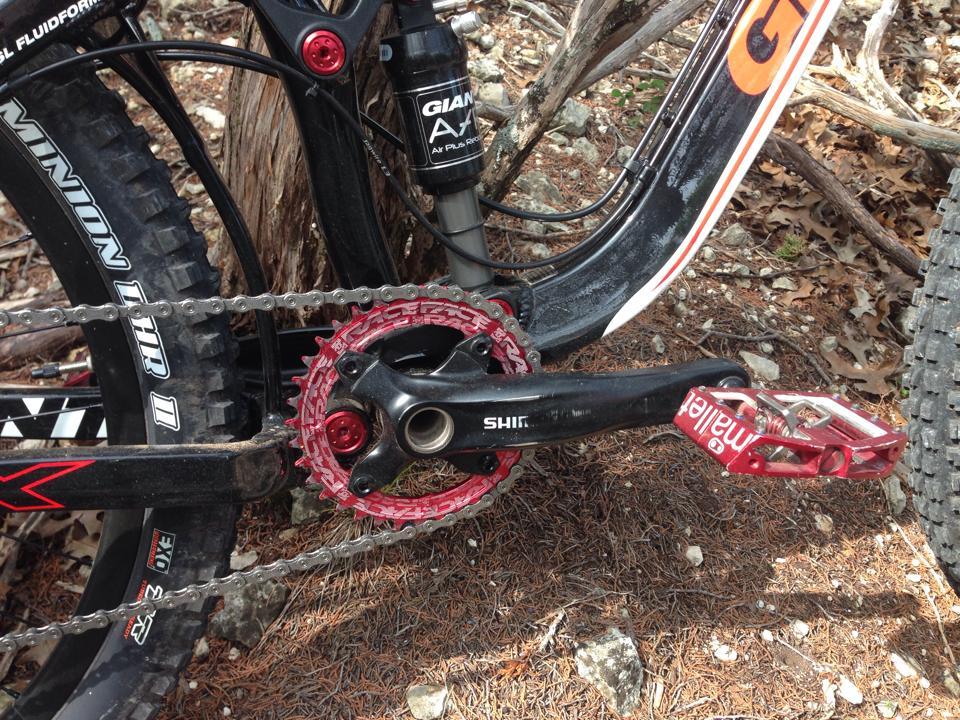 Giant Trance X3: Close-up view of a mountain bike's crankset and pedals, showcasing a black and red color scheme. The image features a chain, a red chainring, and a red pedal with the brand name visible, with a background of pine needles and a tree trunk.