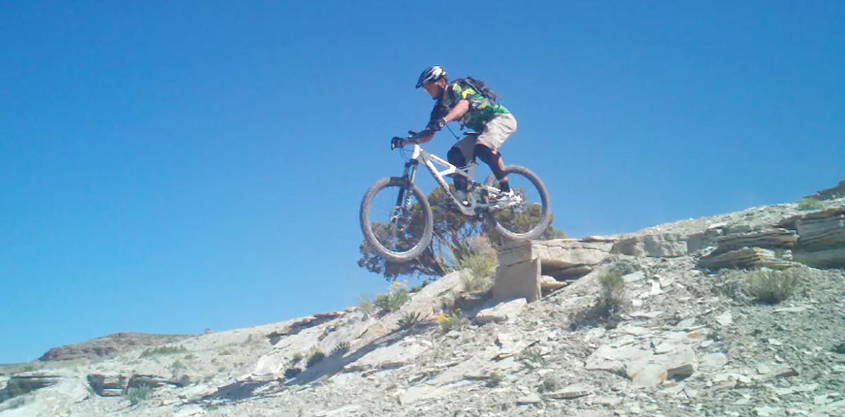 A mountain biker in a green and black shirt is mid-air while jumping off a rocky ledge against a clear blue sky. The terrain is rugged and dry, featuring rock formations and sparse vegetation. Tnt mountain bike trail.