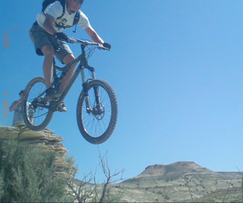 A mountain biker performing a jump off a rocky ledge against a clear blue sky, with a distant mountain in the background. Pick Your Poison mountain bike trail.