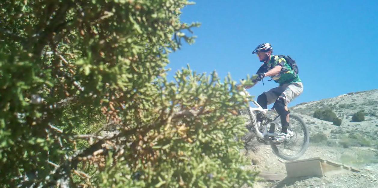A mountain biker performing a jump on a dirt trail, surrounded by greenery and under a clear blue sky. Tnt mountain bike trail.
