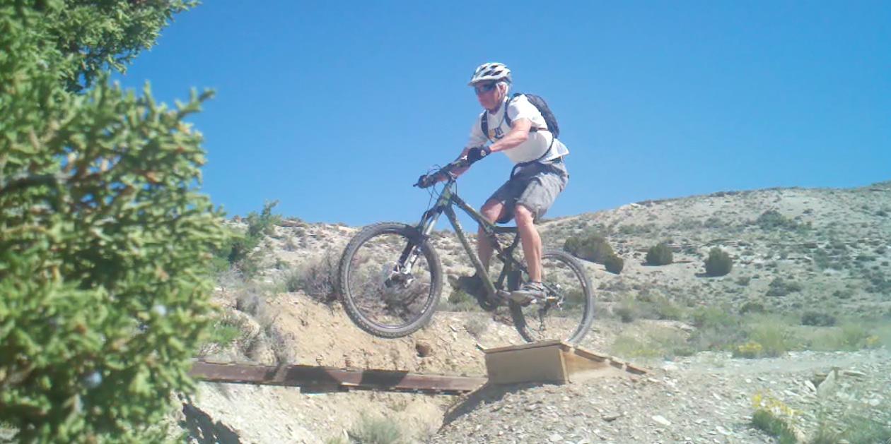 A person in a helmet and sunglasses jumps off a small ramp on a mountain bike, with dirt and shrubs in the background under a clear blue sky. Tnt mountain bike trail.