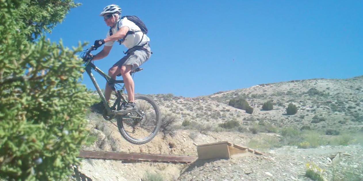 A mountain biker jumps off a small ramp on a rocky terrain, surrounded by shrubs and hills under a clear blue sky. Tnt mountain bike trail.