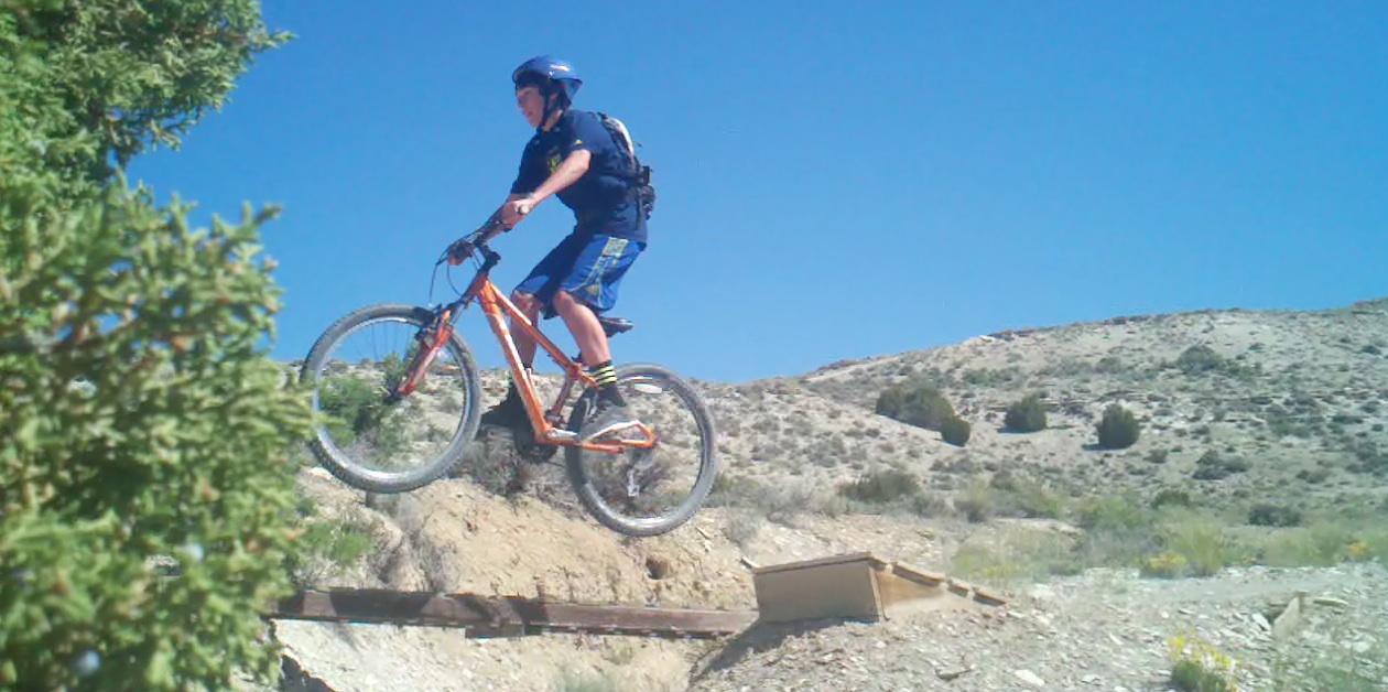 A young person wearing a helmet and biking gear is mid-air on a mountain bike, jumping over a small ramp on a rugged trail surrounded by desert terrain and blue skies. Tnt mountain bike trail.