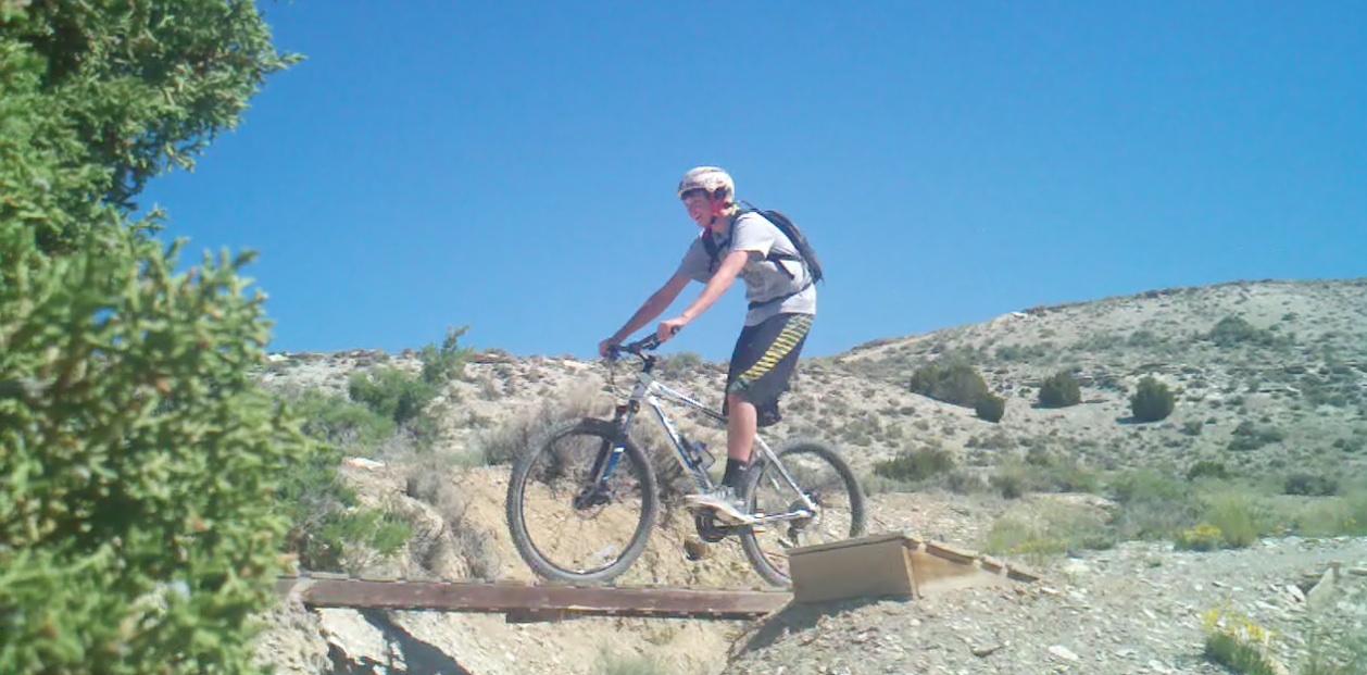 A person riding a mountain bike over a wooden ramp on a dirt trail, surrounded by dry hills and clear blue skies. The rider is wearing a helmet and a backpack, and is captured in motion, leaning forward as they navigate the terrain. Green shrubbery can be seen in the foreground. Tnt mountain bike trail.