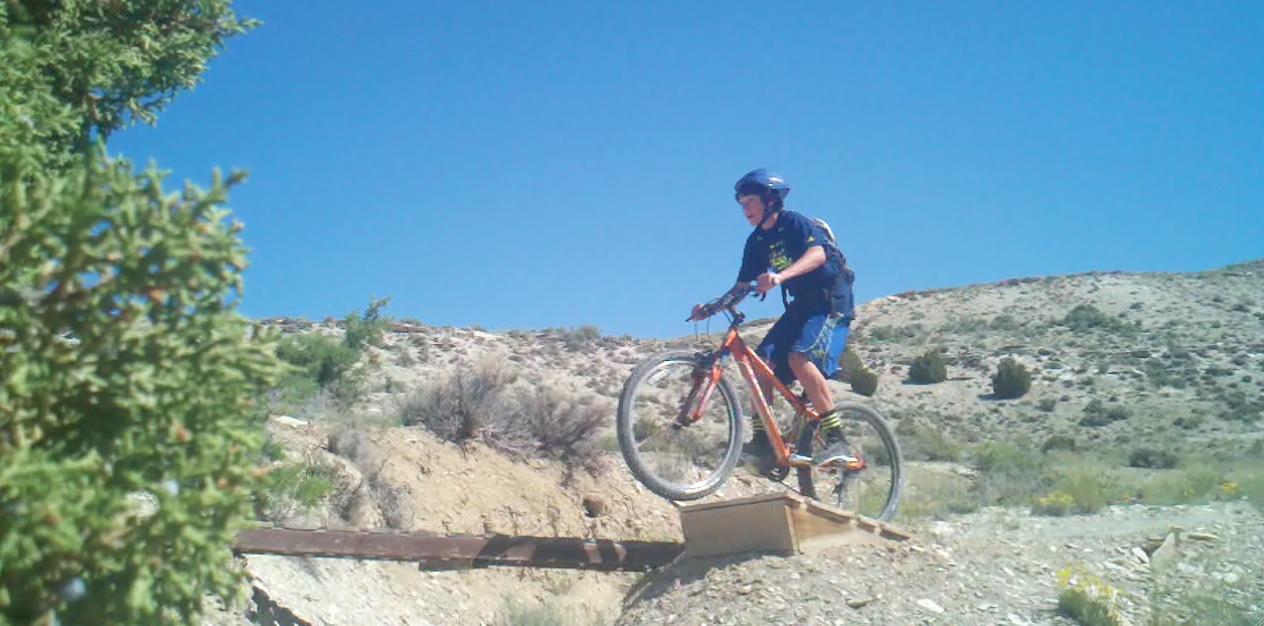 A young cyclist wearing a helmet and casual clothing is jumping over a wooden bridge on a mountain bike. The background features a clear blue sky and rugged terrain with sparse vegetation and hills. Tnt mountain bike trail.