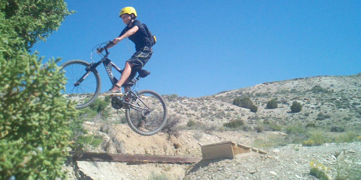 A young person wearing a yellow helmet jumps off a small wooden ramp on a mountain bike, airborne against a clear blue sky, with a mountainous landscape in the background. Tnt mountain bike trail.