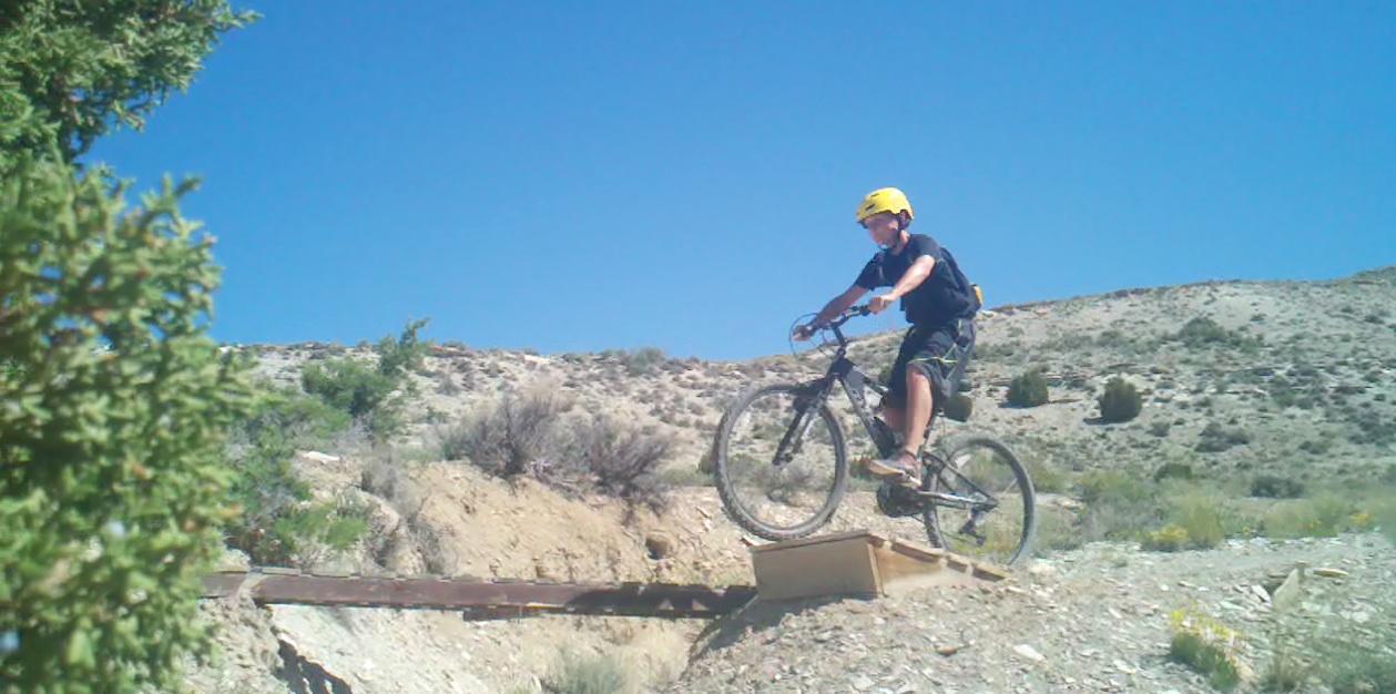 A child in a yellow helmet performs a leap on a mountain bike over a wooden ramp in a desert landscape under a clear blue sky. Tnt mountain bike trail.