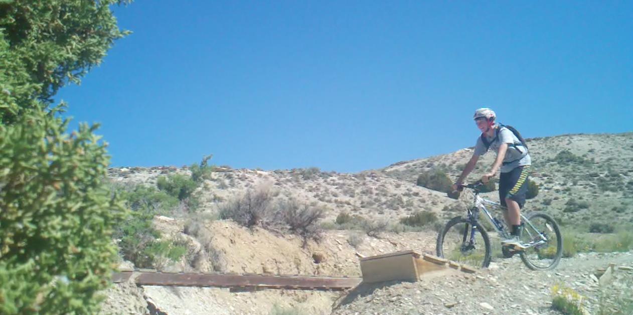 A young man riding a mountain bike on a rugged trail with hills and shrubs in the background, under a clear blue sky. Tnt mountain bike trail.