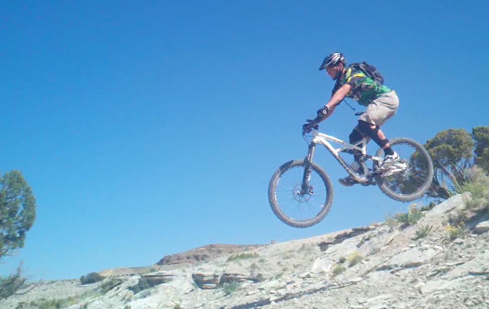 Mountain biker performing a jump on rough terrain under a clear blue sky. The rider is wearing a helmet, protective gear, and a colorful shirt, captured mid-air with the bike wheels off the ground. Tnt mountain bike trail.
