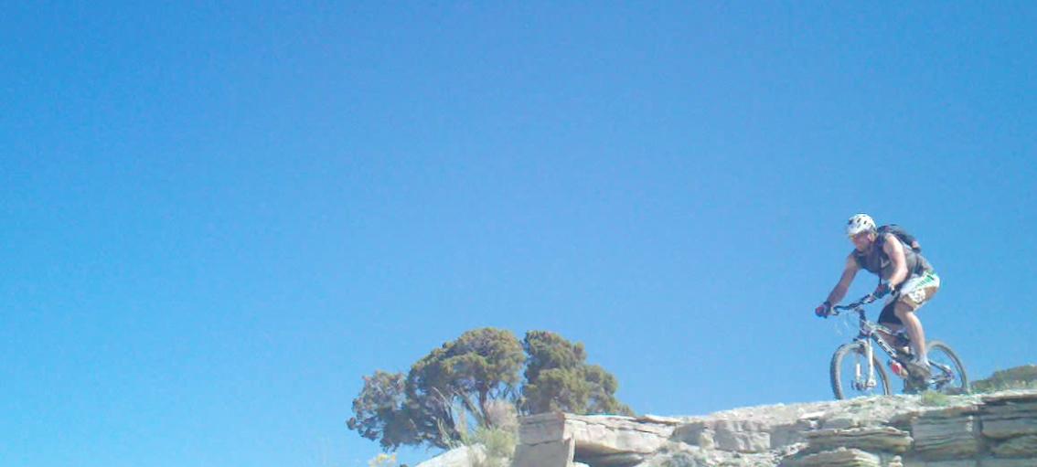 A person riding a mountain bike on rocky terrain under a clear blue sky, with a small tree in the background. Tnt mountain bike trail.