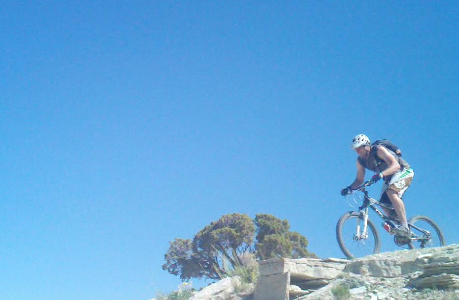 A mountain biker riding on a rocky trail against a clear blue sky, with a small tree in the background. The rider is wearing a helmet and a backpack, showcasing an adventurous outdoor activity. Tnt mountain bike trail.