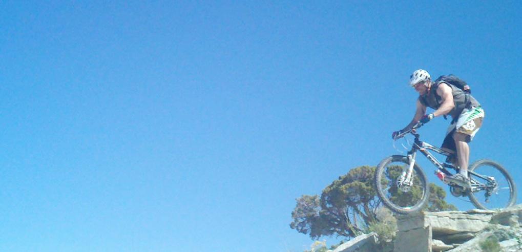 A mountain biker mid-jump over a rock formation against a clear blue sky. The rider is wearing a helmet and a backpack, demonstrating an adventurous spirit in an outdoor setting. Tnt mountain bike trail.
