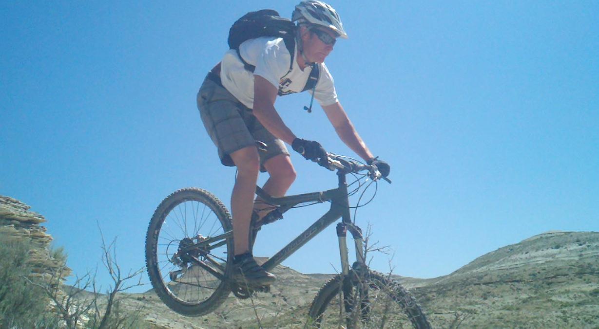 A mountain biker performing a jump while riding on rocky terrain under a clear blue sky. The cyclist is wearing a helmet, sunglasses, a white T-shirt, and gray shorts, with a backpack strapped on. Pick Your Poison mountain bike trail.