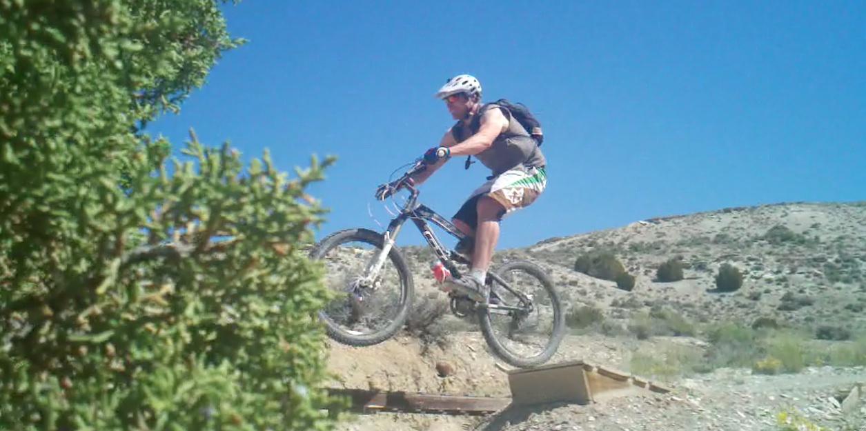 A mountain biker wearing a helmet and sunglasses jumps off a wooden ramp on a dirt trail, surrounded by greenery and against a clear blue sky. Tnt mountain bike trail.