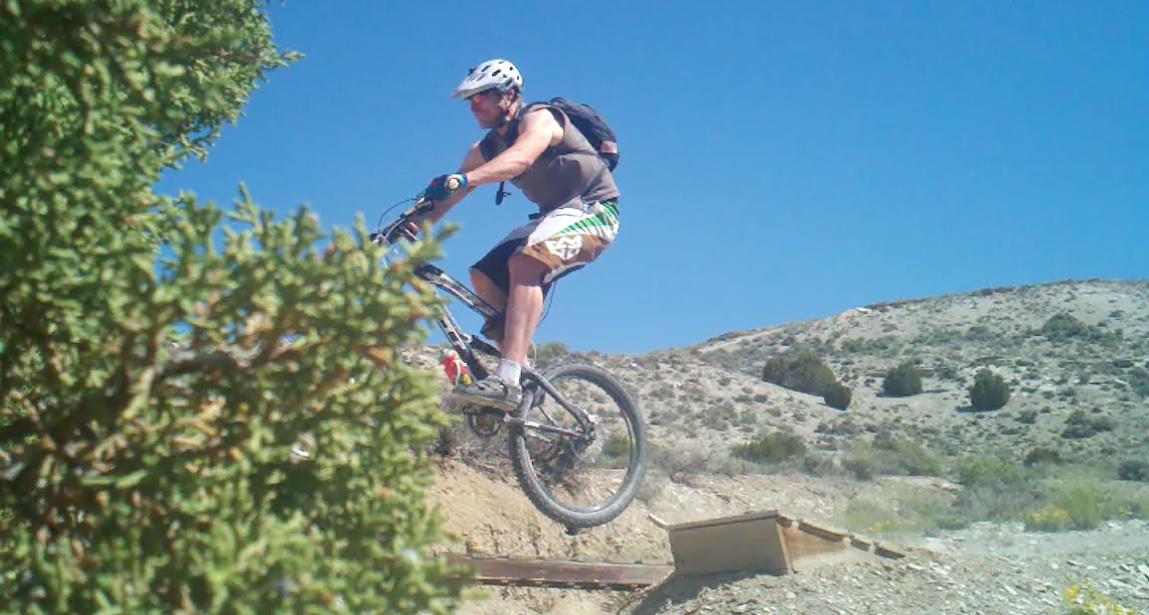 A mountain biker in mid-air jumps off a small wooden ramp on a dirt trail with a clear blue sky in the background. The scene is surrounded by sparse vegetation and rugged terrain, showcasing an outdoor adventure setting. Tnt mountain bike trail.