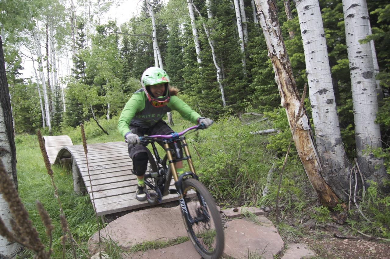 A mountain biker wearing a green jersey and helmet rides swiftly over a wooden bridge on a trail surrounded by lush greenery and tall trees. The scene captures the excitement of mountain biking in a forested area, showcasing the rider's skill and the natural beauty of the outdoors. The Canyons Area mountain bike trail.