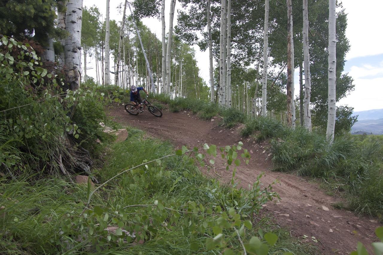A mountain biker leaning into a turn on a dirt trail surrounded by tall aspen trees and green grass, with a cloudy sky in the background. The Canyons Area mountain bike trail.