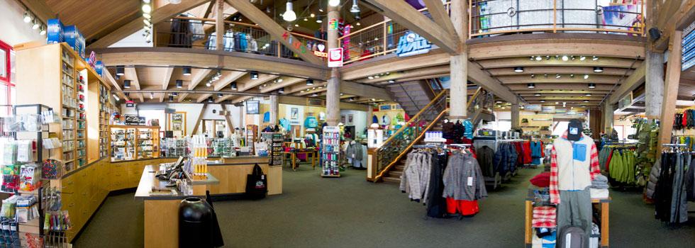 A spacious outdoor retail store interior showcasing various merchandise displays, including clothing and equipment for outdoor activities. The store features wooden beams, a two-level design with a staircase, and an organized layout with shelves and racks filled with jackets, accessories, and outdoor gear. Bright lighting enhances the inviting atmosphere.