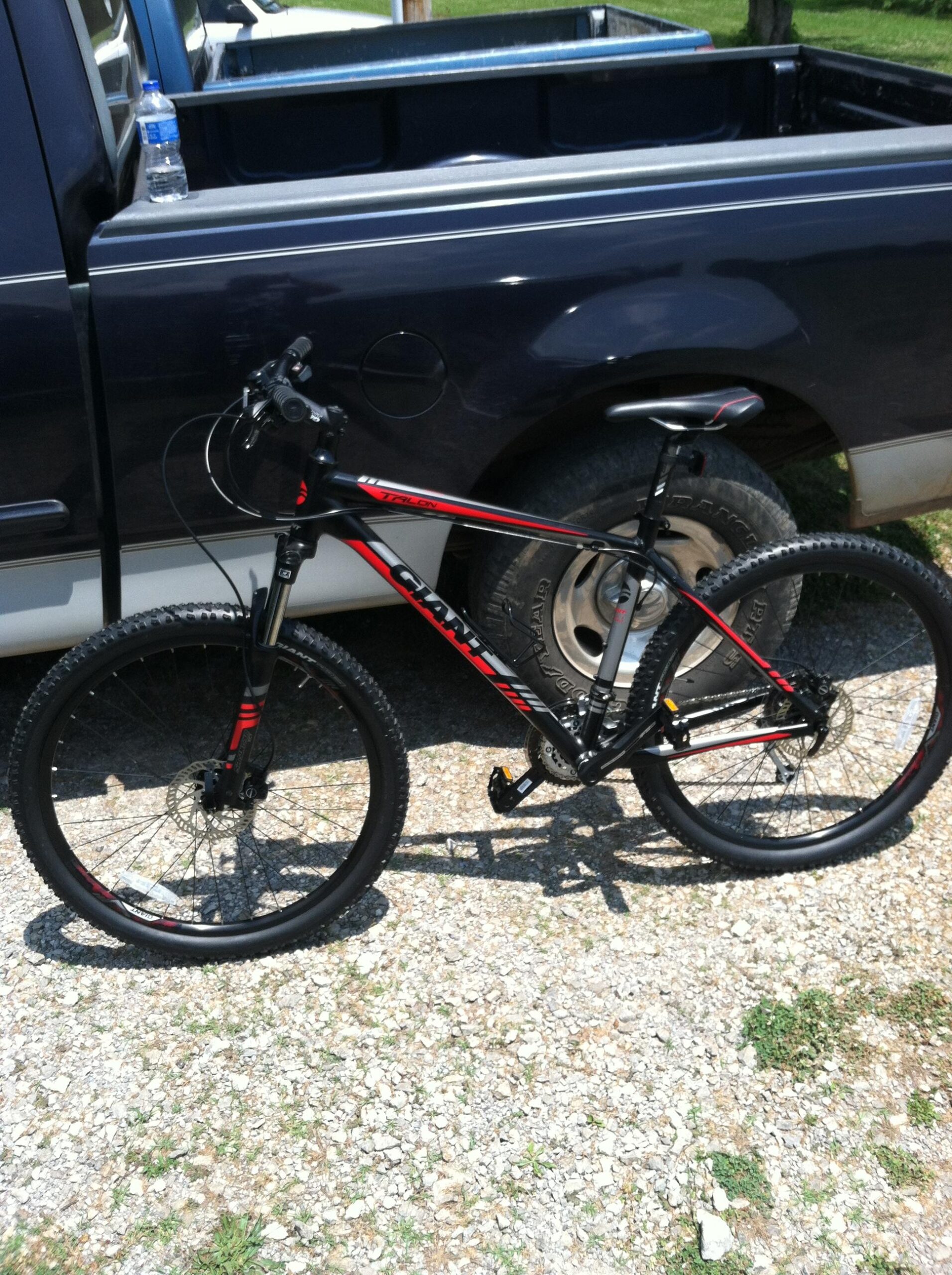 Giant Talon 27.5: A black and red mountain bike is parked beside a pickup truck on a gravel surface. The bike features knobby tires and a sleek frame design. A water bottle is placed on the truck bed in the background.