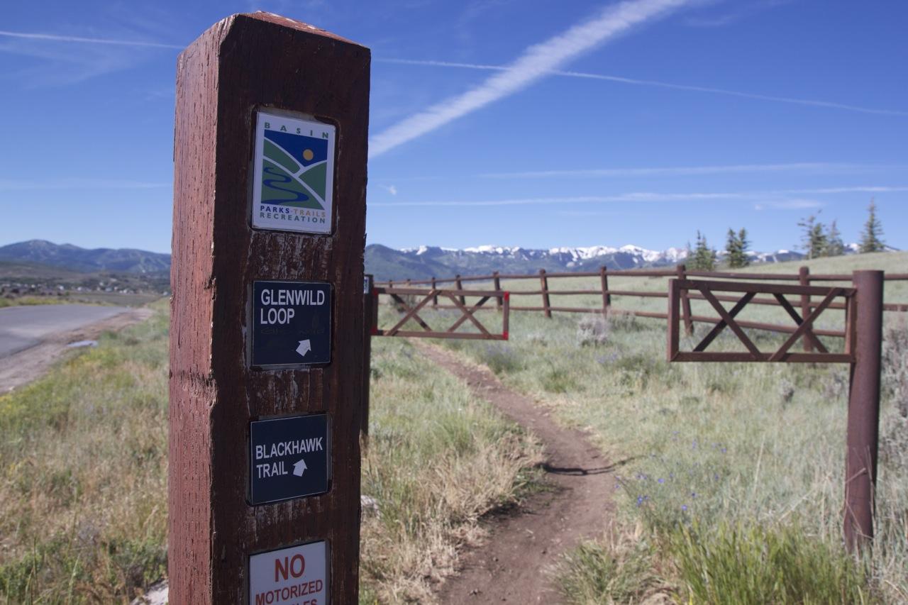 A wooden trail sign post with directional markers for the Glenwild Loop and Blackhawk Trail, situated near a gate that leads into a grassy area with distant mountain views. The sky is clear with a few wispy clouds. A "No Motorized Vehicles" sign is visible at the base of the post. Glenwild mountain bike trail.