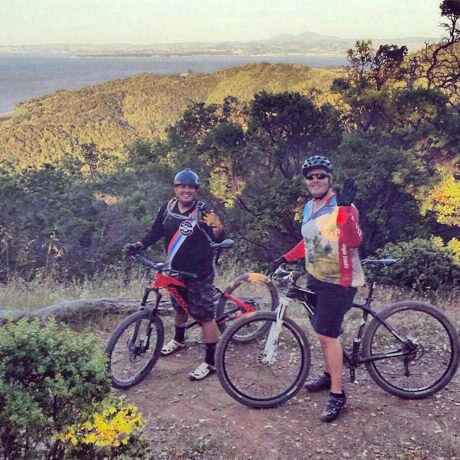 Knolly Chilcotin: Two mountain bikers smiling and waving at the camera while standing beside their bikes on a trail surrounded by greenery. In the background, there are hills and a body of water, indicating a scenic outdoor location.