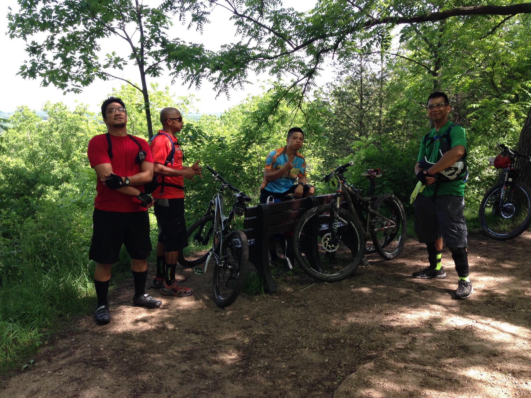 Group of four mountain bikers resting at a trailhead surrounded by lush greenery. They are wearing cycling gear, with two men in red shirts, one in a green shirt, and another in a colorful cycling jersey. Several bicycles are parked nearby, and the atmosphere is relaxed and casual. Emma Carlin mountain bike trail.