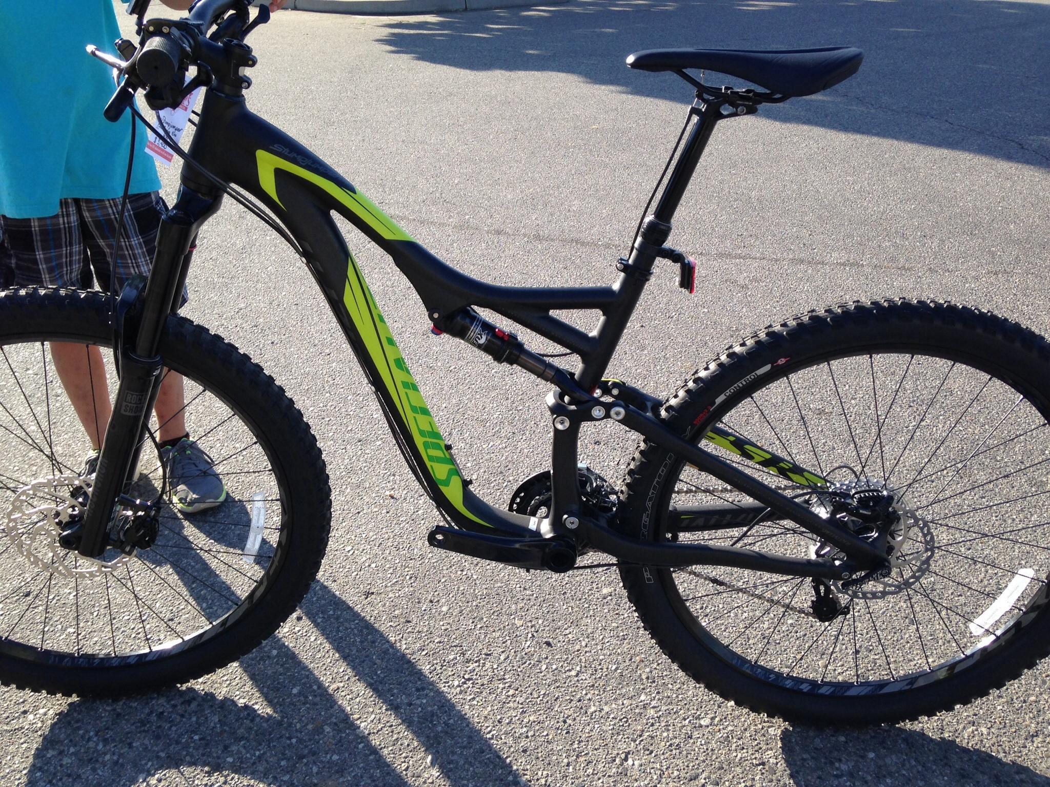 Specialized Stumpjumper FSR Comp EVO: A close-up view of a black and lime green mountain bike, showcasing its frame, handlebars, and wheels. The bike is positioned on a paved surface, with a person in the background holding the handlebars. The bike features thick tires suitable for off-road terrain and a dropper seat post.