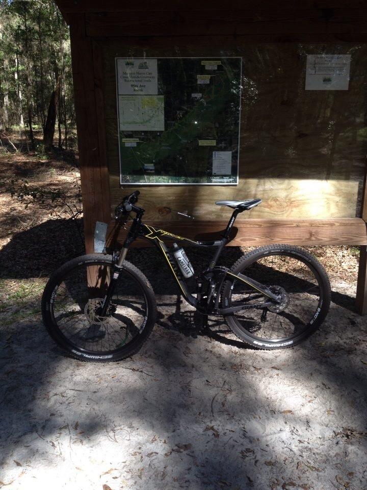 Giant Trance X 29er: A mountain bike parked on a sandy path in front of a wooden information board displaying a map and trail information, surrounded by a forested area with trees and dappled sunlight.