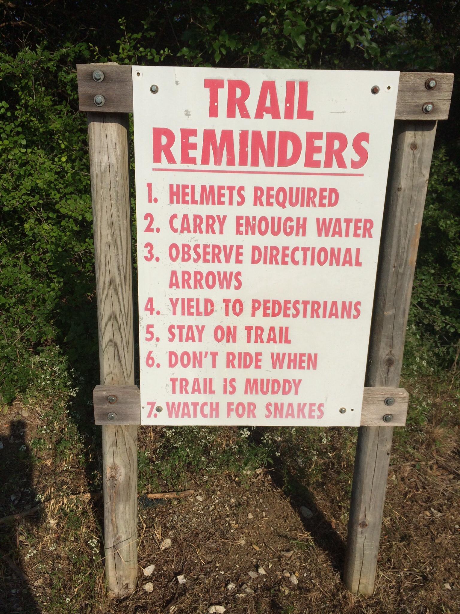 A wooden sign displaying trail reminders with seven safety rules, including "Helmets Required," "Carry Enough Water," and "Watch for Snakes," set against a backdrop of greenery. Cedar Hill State Park At Joe Pool Lake mountain bike trail.