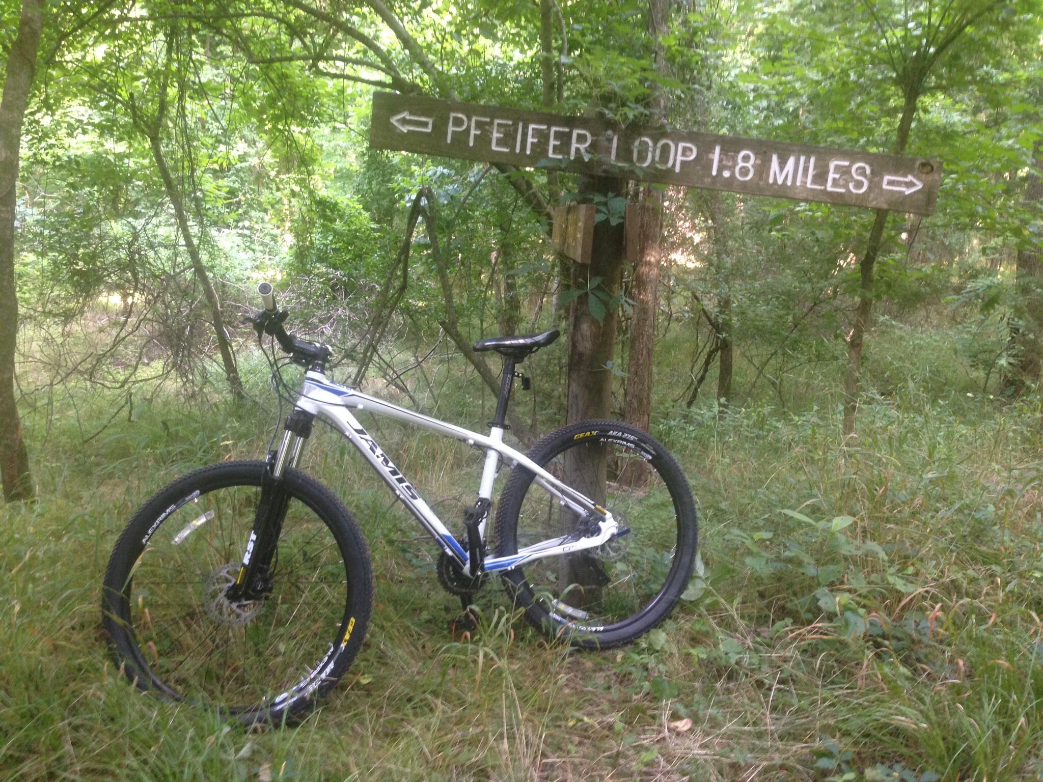 Jamis Nemesis: A mountain bike leaning against a wooden signpost in a lush green forest. The sign indicates the direction and distance to "Pfeifer Loop," which is 1.8 miles away. Surrounding vegetation includes trees and underbrush, creating a peaceful natural setting.