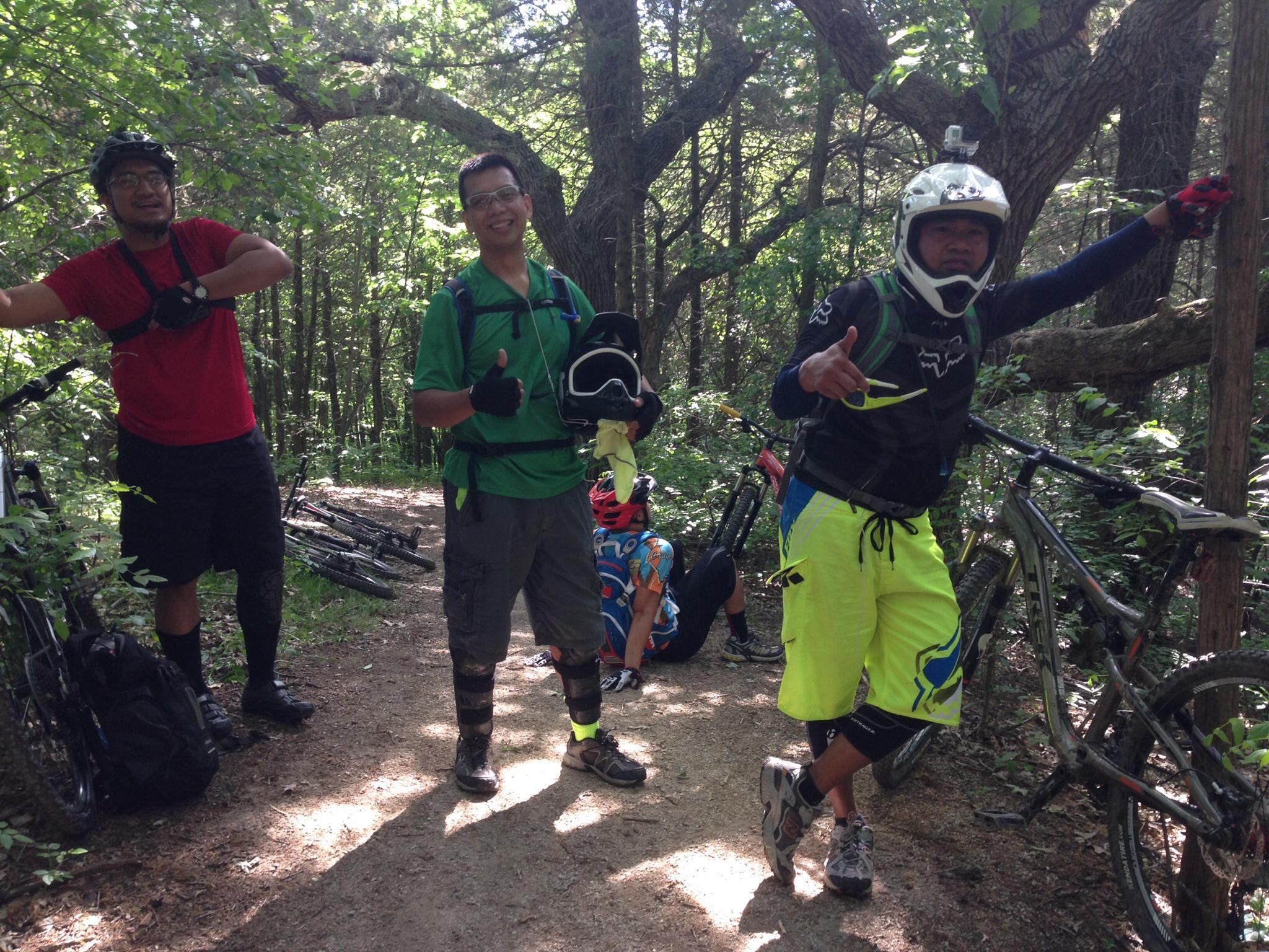 Four mountain bikers pose on a dirt trail surrounded by greenery. Two of them are standing, smiling, and wearing helmets and biking gear, while one is leaning against a tree and giving a thumbs up. The fourth biker is sitting on the ground in the background. Several mountain bikes are parked nearby. The scene captures a fun and active moment in nature. Emma Carlin mountain bike trail.