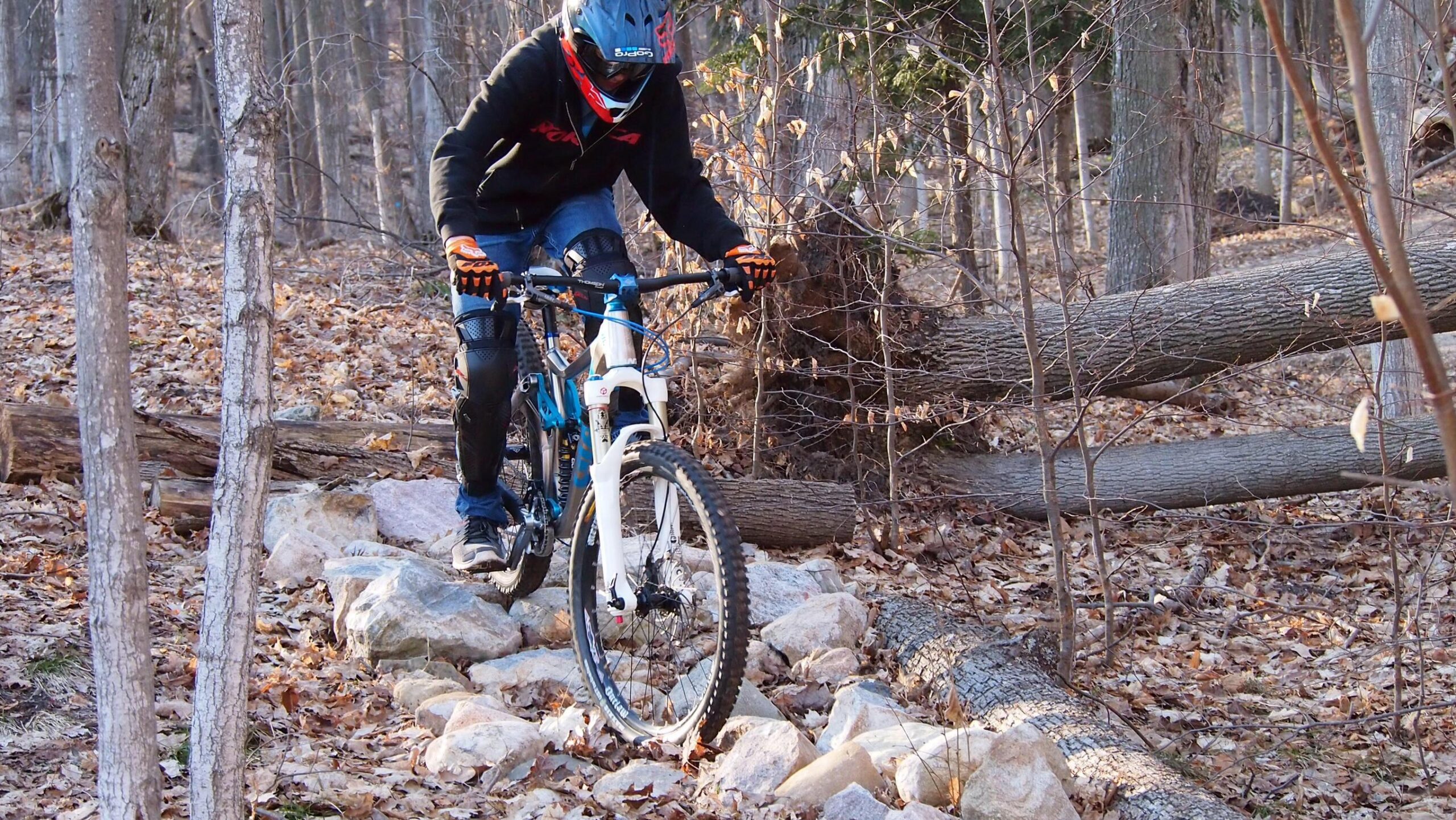 A mountain biker navigating over rocks and fallen logs on a forest trail, surrounded by trees and dried leaves. The rider is wearing protective gear, including a helmet and gloves, and is focused on maintaining balance as they maneuver through the rugged terrain. Copper Ridge Freeride mountain bike trail.