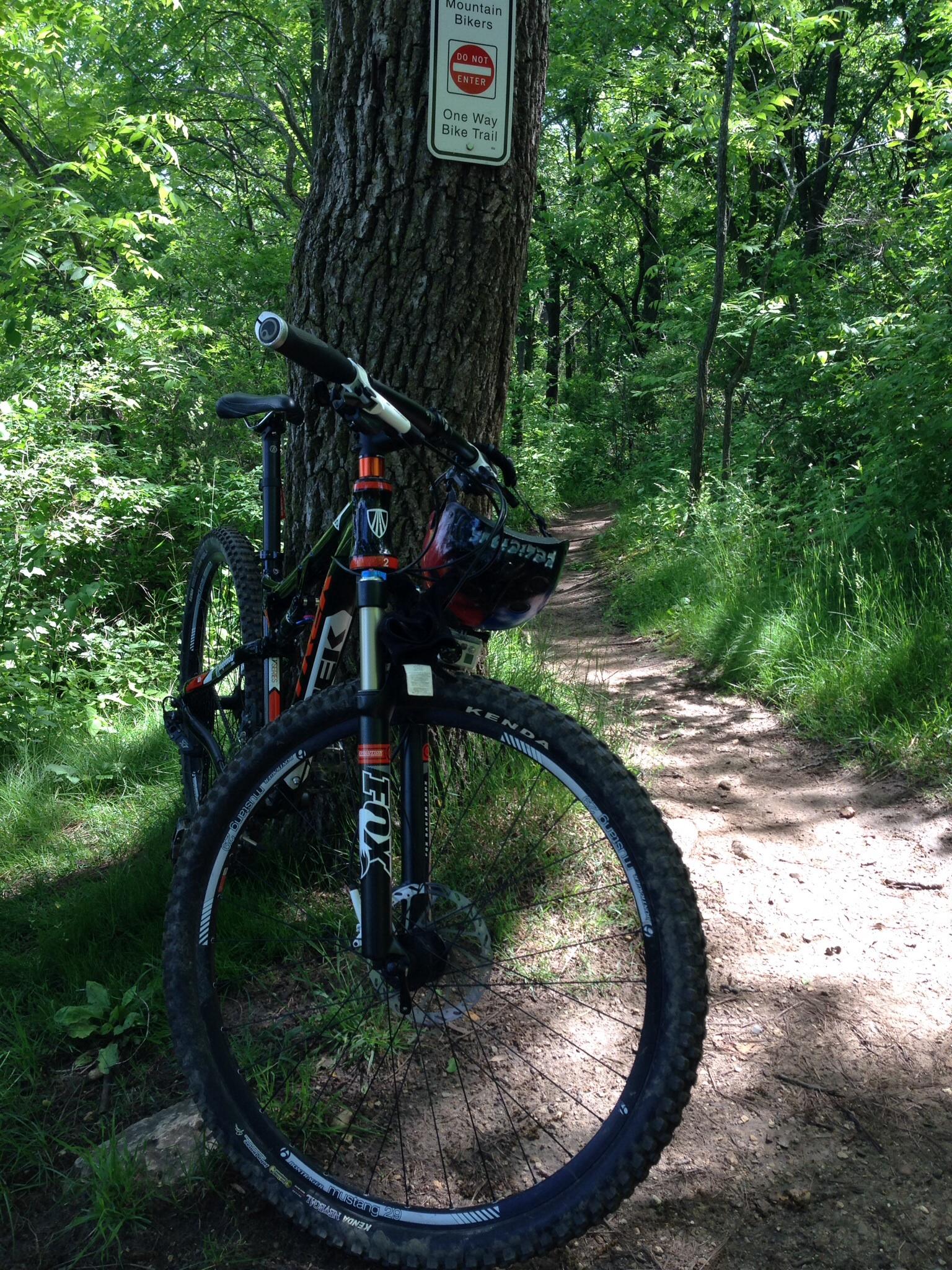 A mountain bike leaned against a tree on a narrow dirt trail surrounded by lush greenery. A sign on the tree indicates a one-way bike trail with a "Do Not Enter" warning for mountain bikers. Emma Carlin mountain bike trail.