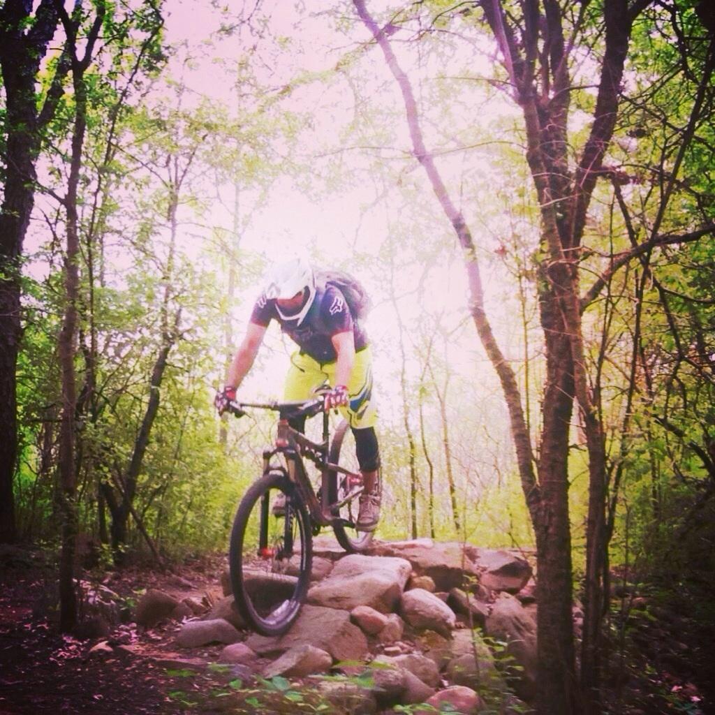 A mountain biker dressed in bright yellow gear is jumping over a rocky terrain in a wooded area. The sunlight filters through the trees, creating a vibrant and lively atmosphere. Levis Mounds mountain bike trail.